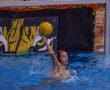 Water polo player prepares to throw the ball during a competitive match near the goal, tiger mascot in background.