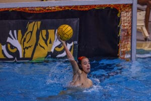 Water polo player prepares to throw the ball during a competitive match near the goal, tiger mascot in background.