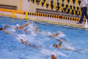 Water polo players competing in an action-packed match in an indoor pool with referee watching.