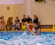 Water polo player in action, throwing a ball across the pool during a game. Spectators seated in the background.