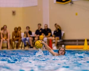 Water polo player in action, throwing a ball across the pool during a game. Spectators seated in the background.