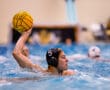 Water polo player in action wearing a black cap, poised to throw a yellow ball in a competitive indoor match.