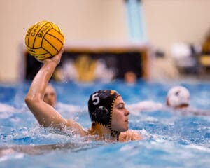 Water polo player in action wearing a black cap, poised to throw a yellow ball in a competitive indoor match.