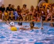 Water polo players in intense game action with yellow ball, poolside spectators in background, splashing water around.