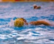 Water polo player in action, swimming with a yellow ball on a vibrant blue pool background.