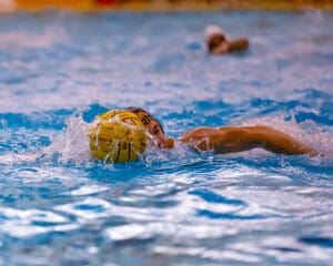 Water polo player in action, swimming with a yellow ball on a vibrant blue pool background.