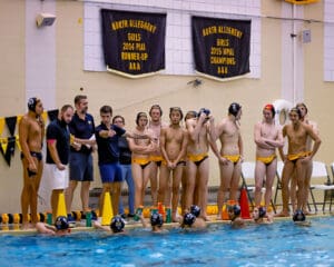 Water polo team strategizes poolside, coach giving instructions, preparing for upcoming game. Championship banners in background.