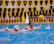 Water polo player in action, holding the ball high, while a competitor approaches in a pool with colorful banners.