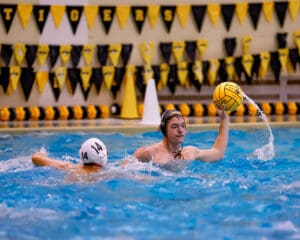 Water polo player in action, holding the ball high, while a competitor approaches in a pool with colorful banners.
