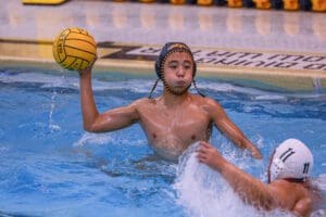 Water polo player prepares to throw ball during intense match in pool, wearing cap, surrounded by water splashes.