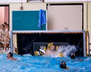 Water polo goalie blocks shot as players converge in swimming pool match.