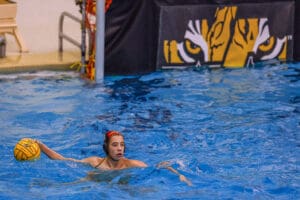 Water polo player in blue pool holding yellow ball, tiger logo in background.