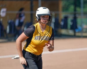 Softball player in action, wearing a helmet and yellow jersey, ready to sprint during a game on a sunny day.