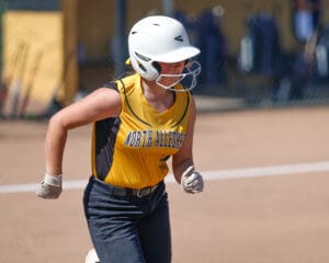 Softball player in yellow jersey runs on the field during a game, wearing a white helmet and gloves.