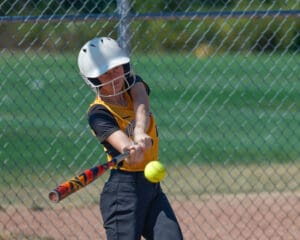 Softball player in yellow jersey hits ball with a colorful bat on a sunny day, chain-link fence in background.