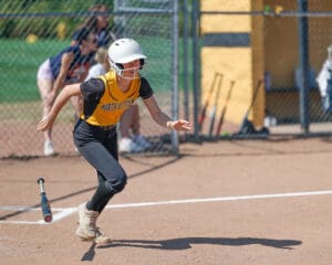 Softball player in a yellow jersey runs from home plate, bat discarded, during a game on a sunny day.