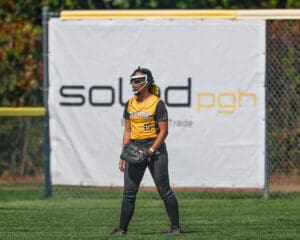 Softball player in yellow uniform stands ready on the field near a fence with a branded banner.