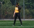 Softball player in yellow jersey and black pants stands ready on the field, glove in hand, with a fence in the background.