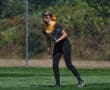 Softball player in yellow and black uniform prepares to field a ball on a grassy field with a fence background.