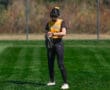 Softball player in a yellow jersey and helmet stands on a green field, adjusting her gloves during a sunny day.