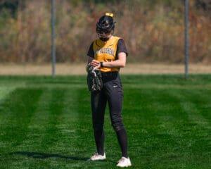 Softball player in a yellow jersey and helmet stands on a green field, adjusting her gloves during a sunny day.