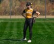 Softball player in yellow and black uniform standing on green field, wearing helmet and glove, ready for action.