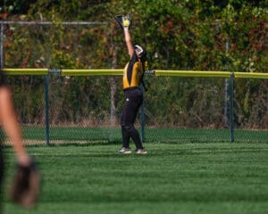 Softball player catches fly ball at outfield fence in mid-game action.