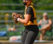 Softball player in action wearing a helmet and visor, poised to throw with glove on, blurred spectators in background.