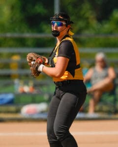 Softball player in action wearing a helmet and visor, poised to throw with glove on, blurred spectators in background.