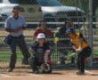 Softball player hits a pitch during a game with umpire and catcher visible. Focused athletic action shot.