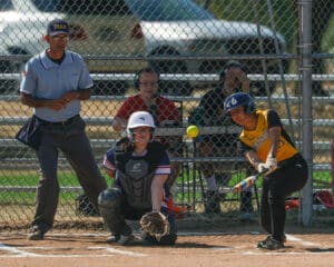 Softball player hits a pitch during a game with umpire and catcher visible. Focused athletic action shot.