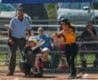 Batter hits softball as umpire and catcher watch during daytime game, chain-link fence in background.