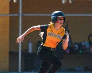 Softball player in action wearing a helmet and yellow jersey, North Allegheny team, sprinting on the field.