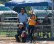 Softball player hits the ball during a game, with an umpire and catcher behind, under a sunny sky.