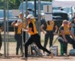 Softball player running past teammates in dugout during a game, wearing black and yellow uniform and white helmet.
