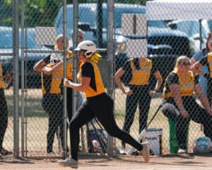 Softball player running past teammates in dugout during a game, wearing black and yellow uniform and white helmet.