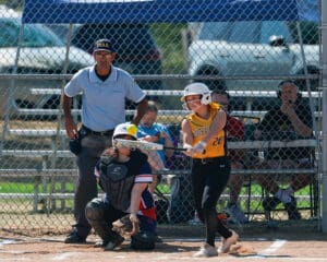 Softball batter swings at a pitch during a game, with an umpire and catcher in position behind the plate.