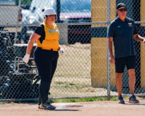Softball player in yellow jersey at base with coach nearby, chain-link fence in background.