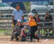 Softball player hits a fastpitch during a game, umpire and catcher in position, spectators in background.