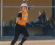 Softball player running bases in full gear during a game with dugout in background.