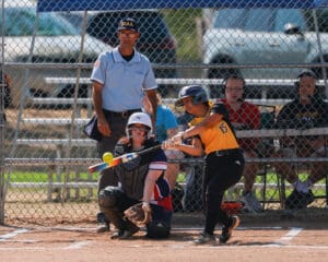 Softball player in action swinging at a pitch, with a catcher and umpire behind at a sunny outdoor game.