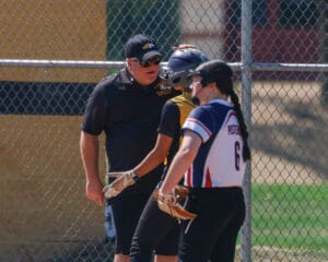Softball coach offers guidance to players beside a fenced field during a sunny day game.