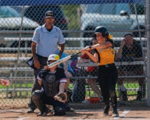 A softball player in a yellow jersey swings at a pitch, with a catcher and umpire in the background on a sunny day.