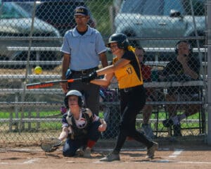 Softball player in yellow jersey batting, catcher ready to catch, during a game at a fenced field.