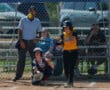 Softball player swinging bat during game with umpire and catcher, sunny field, action shot.