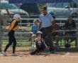 Youth softball game: player batting, catcher crouching, umpire observing, chain-link fence backdrop, summer setting.