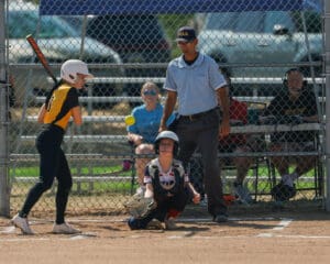 Youth softball game: player batting, catcher crouching, umpire observing, chain-link fence backdrop, summer setting.