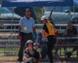 Softball player in yellow jersey prepares to bat as umpire and catcher watch the approaching pitch.