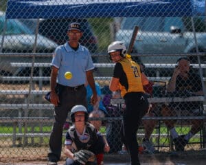 Softball player in yellow jersey prepares to bat as umpire and catcher watch the approaching pitch.