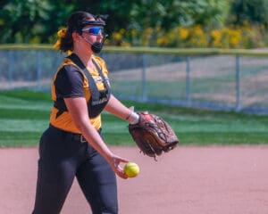 Softball player in action on field, wearing safety gear and black-and-yellow uniform, holding ball and glove.